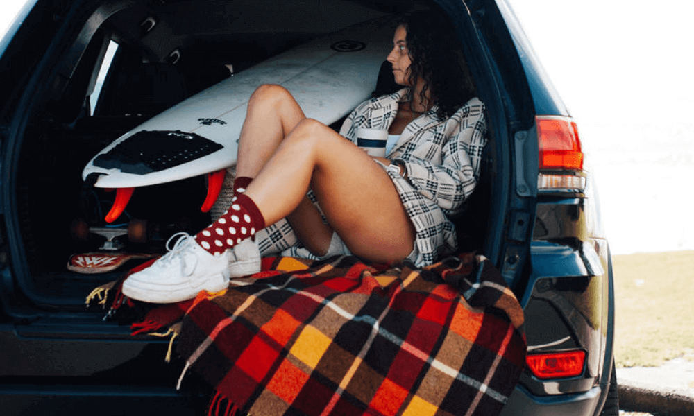 Woman wearing merino socks for travel sitting in a car with a surfboard, enjoying a scenic view.