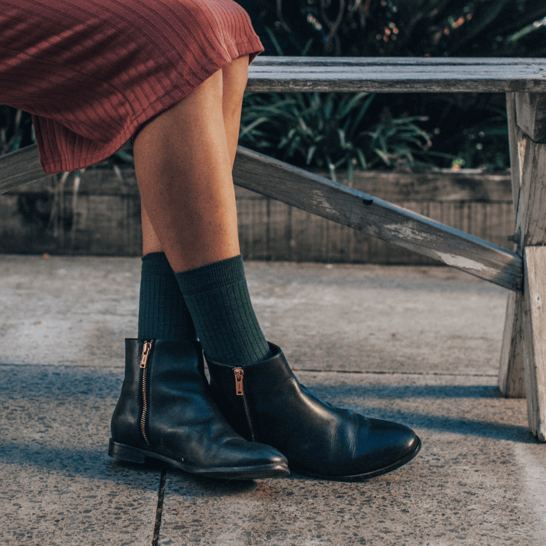Woman wearing Merino Wool Crew Socks in green with black ankle boots, sitting on a bench.